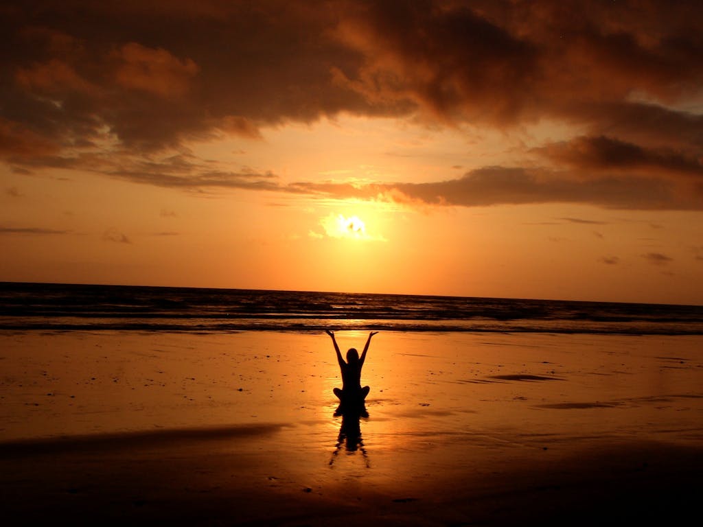 Silhouette of Person Raising Its Hand above a sun while sitting on the shoreline of a beach