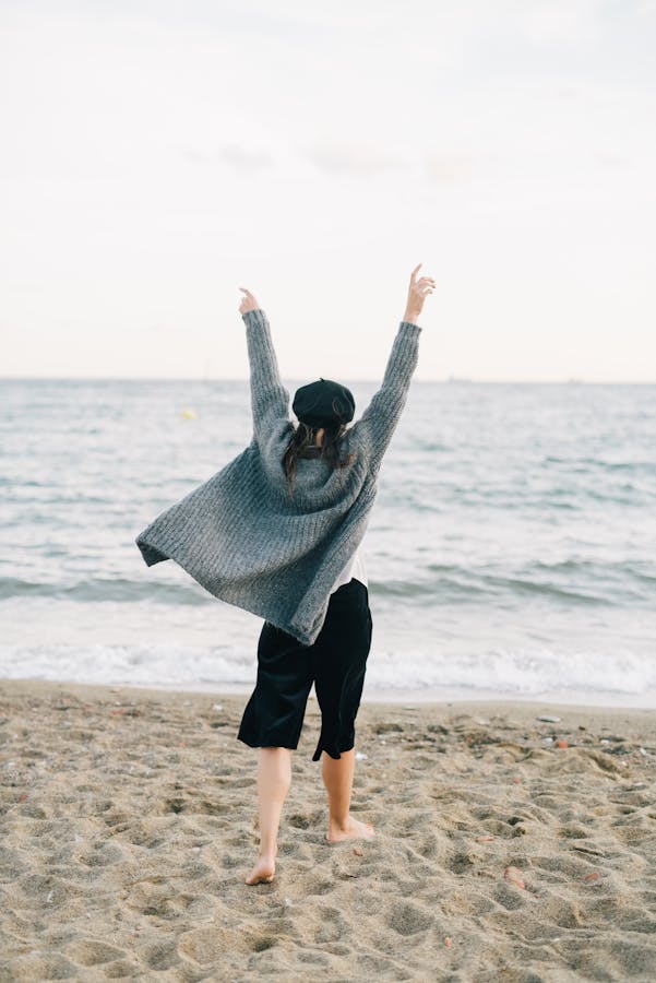 A Person in Gray Cardigan Walking on the Shore with Arms Raised